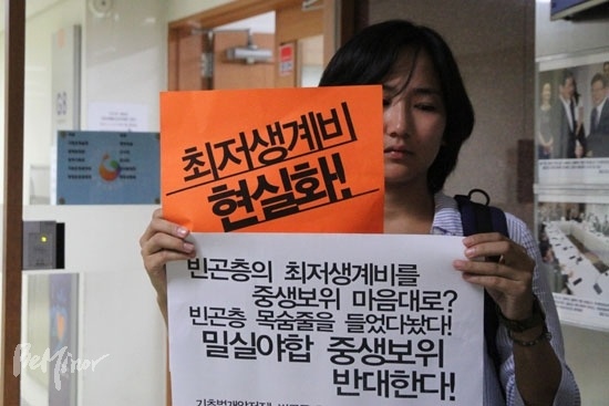 2014년 빈곤단체 활동가가 중생보위 회의장 앞에서 "빈곤층의 최저생계비를 중생보위 마음대로?" 등의 문구가 적힌 피켓을 들고 1인 시위를 하는 모습. 문진영 교수는 최저생계비가 중위소득으로 대체되면서 급여 기준선을 정부 재량으로 결정하는 것이 더 쉬워졌다고 지적했다.