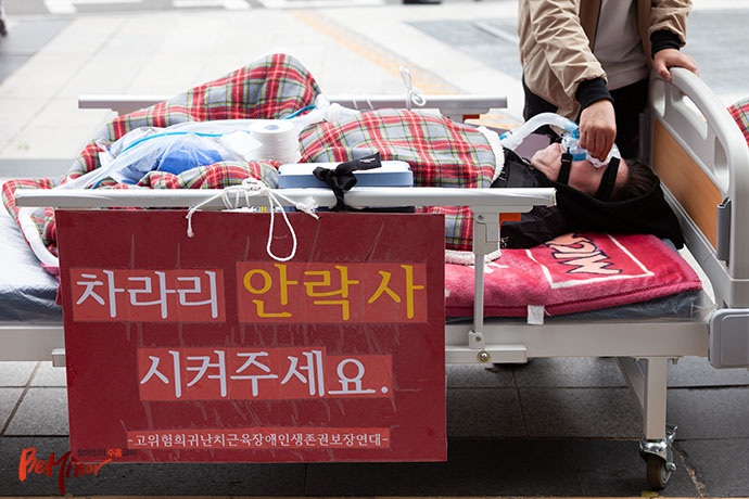 인공호흡기를 착용한 채 침대에 누워있는 근육장애인 배현우 씨. 침대엔 “차라리 안락사 시켜주세요”라는 문구가 적힌 커다란 팻말이 걸려 있다.