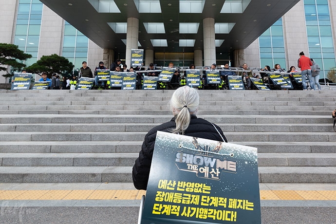 박경석 전국장애인차별철폐연대 상임공동대표가 발언하고 있다. 그 앞으로 국회의원회관 앞에서 쇠사슬을 몸에 감은 채, 점거하고 있는 장애인활동가들의 모습이 보인다. 