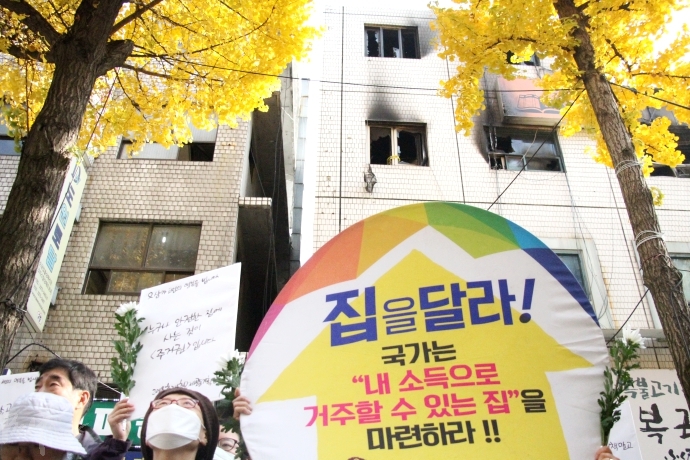 기자회견 참석자들이 들어올린 피켓에 '집을 달라!'는 문구가 적혀있다. 피켓 너머로 화재 피해를 입은 국일고시원 건물이 보인다.
