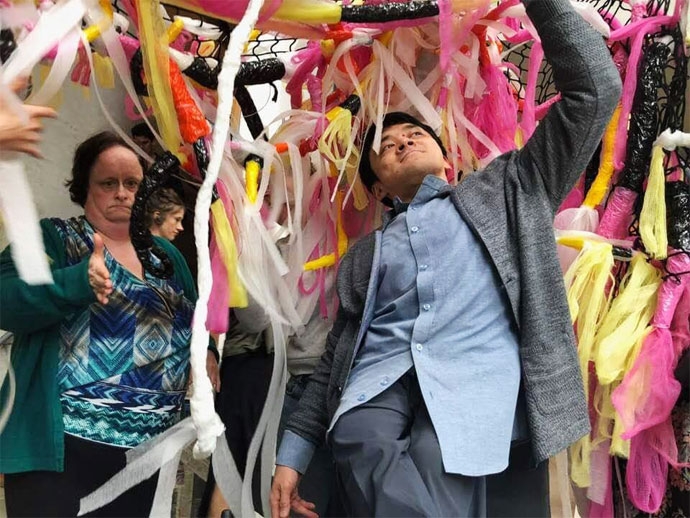 발달장애인 예술가의 작업(흰색, 노란색, 분홍색의 끈들이 아래로 늘어뜨린 설치물) 아래서 필자가 왼팔을 들어 올리고 오른팔은 반대로 내린 채 자세를 뒤로하고 신나있다