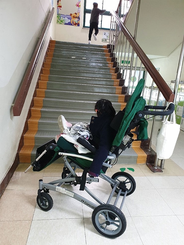 휠체어를 이용하는 지적장애아동 예현이(사진 속 휠체어 탄 사람)는 올해 3월 일반학교 특수학급에 입학했다. 그러나 교실은 엘리베이터 없는 건물 2층에 있다. 계단 앞에 멈춰서 있는 예현이의 모습. (사진 제공 : 심정선 씨) 