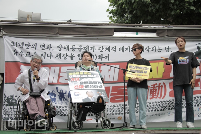 장애등급제 ‘진짜’ 폐지를 위한 투쟁 선포식에서 발언을 하고 있는 변경택 한국뇌병변장애인인권협회 회장, 손팻말을 들고 발언을 기다리고 있는 최용기 한국장애인자립생활센터협의회 회장과 김종옥 전국장애인부모연대 부회장의 모습. 옆에는 수어통역사가 수어통역을 하고 있다. 사진 강혜민 장애등급제 ‘진짜’ 폐지를 위한 투쟁 선포식에서 발언을 하고 있는 변경택 한국뇌병변장애인인권협회 회장, 손팻말을 들고 있는 최용기 한국장애인자립생활센터협의회 회장과 김종옥 전국장애인부모연대 부회장의 모습. 옆에는 수어통역사가 수어통역을 하고 있다. 사진 강혜민