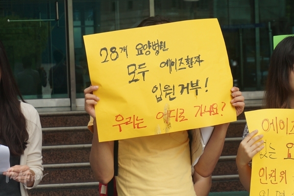 한 사람이 “28개 요양병원 모두 에이즈환자 입원 거부! 우리는 어디로 가나요?”라고 적힌 손피켓을 들고 있다. 한 사람이 “28개 요양병원 모두 에이즈환자 입원 거부! 우리는 어디로 가나요?”라고 적힌 손피켓을 들고 있다.