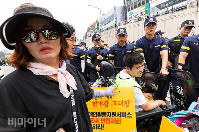 결의대회를 마친 전장연은 오후 4시 더불어민주당 당사가 있는 여의도 방향으로 행진을 했다. 가는 도중 충정로 사거리 육교에서 장애계 요구안이 담긴 대형 현수막을 내 거는 과정에서 경찰과 마찰을 빚기도 했다. 휠체어 뒤에 걸린 손팻말에는 '장애인활동지원서비스 만 65세 연령제한 폐지하라!'라고 적혀있다. 사진 박승원