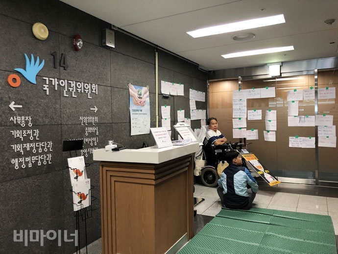 인권위 상임위원회 회의가 열리는 회의실 앞에서 피케팅하는 송용헌 씨. 그의 곁에 활동지원사가 있다. 연령 제한을 의미하는 65가 커다랗게 붙어있다. 사진 강혜민