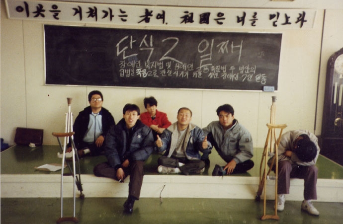 1989년 11월, 최옥란 열사(왼쪽에서 세 번째)가 장애인문제연구회 울림터 회원들과 장애인고용촉진법 제정, 심신장애자복지법 개정 등 양대법안 제·개정을 촉구하며 신민주공화당사를 점거, 단식투쟁을 전개하고 있다. 칠판에는 단식 2일째라고 적혀 있으며, 위에는 “이곳을 거쳐 가는 자여, 조국은 너를 믿노라”라는 문구가 새겨져 있다. 사진제공 빈곤사회연대 1989년 11월, 최옥란 열사(왼쪽에서 세 번째)가 장애인문제연구회 울림터 회원들과 장애인고용촉진법 제정, 심신장애자복지법 개정 등 양대법안 제·개정을 촉구하며 신민주공화당사를 점거, 단식투쟁을 전개하고 있다. 칠판에는 단식 2일째라고 적혀 있으며, 위에는 “이곳을 거쳐 가는 자여, 조국은 너를 믿노라”라는 문구가 새겨져 있다. 사진제공 빈곤사회연대