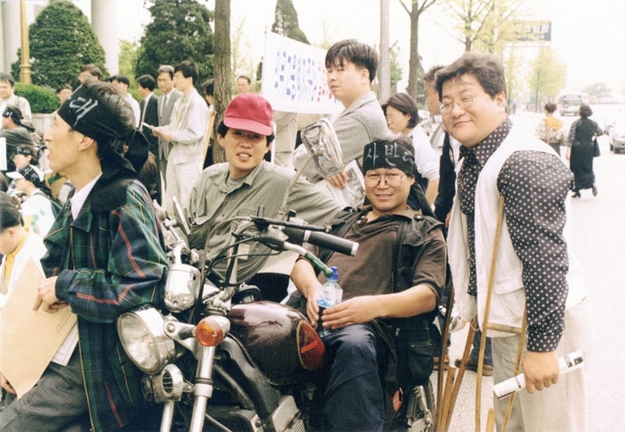 그의 트레이드마크인 삼륜 오토바이를 타고 ‘결사반대’라는 머리띠를 한 박흥수 열사(가운데). 오른쪽은 정태수 열사. 사진 장애해방열사 단 94년, 재계가 2%였던 장애인 의무고용률을 1%로 하향 조정하려 하자 전국장애인한가족협회는 대규모 집회를 열어 강력히 반발한다. 당시 그의 트레이드마크인 삼륜 오토바이를 타고서 ‘결사반대’라는 머리띠를 하고 집회에 참석한 박흥수 열사(가운데). 오른쪽은 정태수 열사. 사진 장애해방열사 단