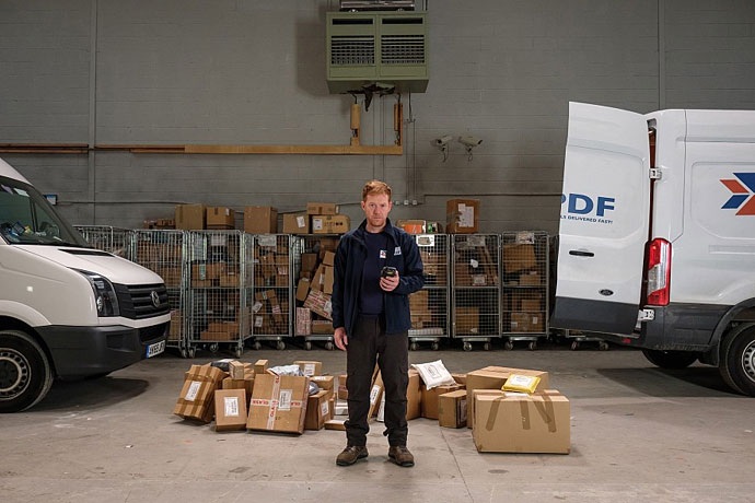 영화 ‘미안해요 리키’ 스틸컷. 리키가 택배공장 안에서 바코드 찍는 기계를 손에 들고 있다. 그의 뒤로 수많은 택배 박스가 쌓여 있으며, 양쪽에는 택배 운송 차량이 세워져 있다. 