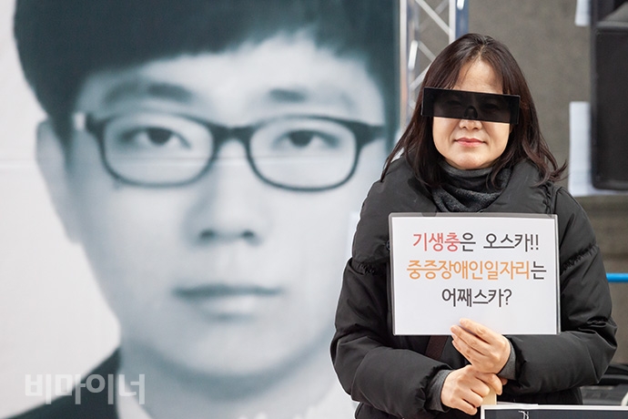 강희석 나야장애인인권교육센터 대표가 눈 모자이크 모양의 선글라스를 쓰고 ‘기생충은 오스카! 중증장애인일자리는 어째스카?’라고 적힌 손팻말을 들고 있다. 그 뒤로는 고 설요한 영정사진이 보인다. 사진 박승원