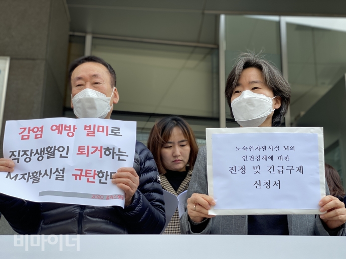 한 활동가가 ‘감염 예방 빌미로 직장생활인 퇴거하는 M자활시설 규탄한다’라고 적혀있는 피켓을 들고 있으며, 그 오른편에는 진정인들의 대리인을 맡은 장서연 공익인권법재단 공감 변호사가 ‘노숙인자활시설 M의 인권침해에 대한 진정 및 긴급구제 신청서’를 들고 있다. 사진 이가연