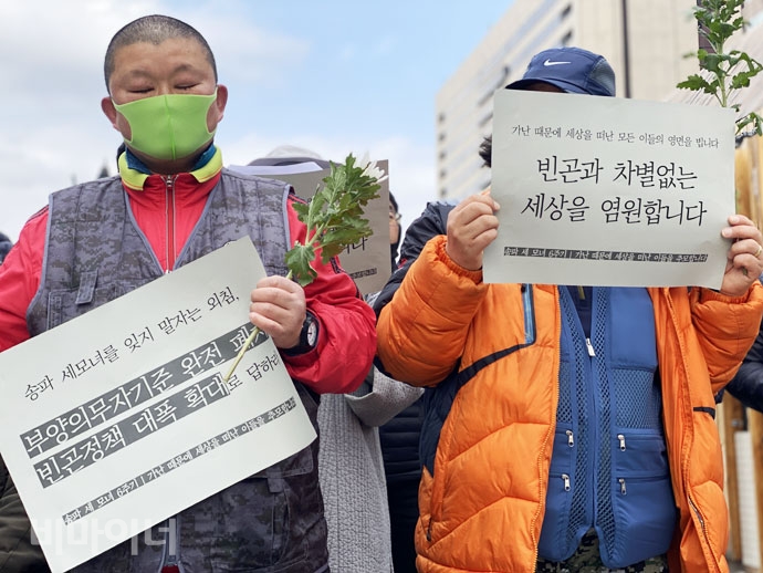 2월 26일, 기초법바로세우기공동행동 등이 개최한  ‘송파 세 모녀 6주기 및 가난 때문에 세상을 떠난 이들의 추모 기자회견’에서 한 참가자가 국화꽃과 피켓을 들고 있다. 피켓에는 ‘송파 세모녀를 잊지 말자는 외침, 부양의무자기준 완전 폐지, 빈곤정책 대폭 확대로 답하라’, ‘빈곤과 차별없는 세상을 염원합니다’라고 적혀 있다. 사진 이가연