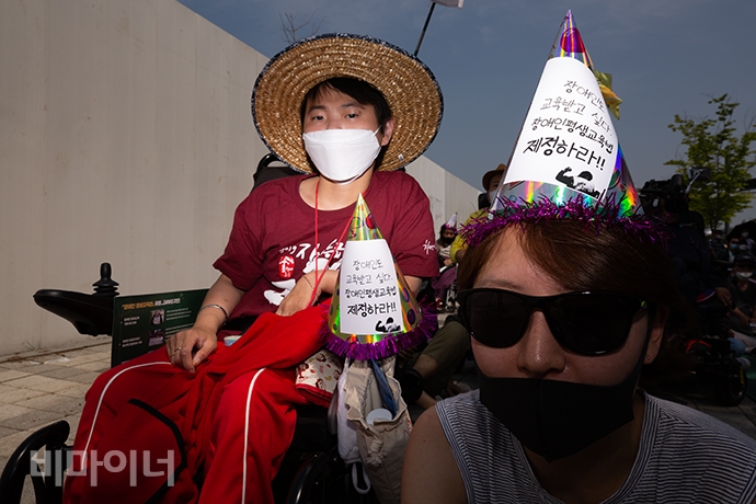 이혜미 노들장애인야간학교 학생(왼쪽부터)과 박임당 노들장애인야간학교 교사가 함께 진행 중인 결의대회를 보고 있다. 고깔모자에는 ‘장애인도 교육받고 싶다. 장애인평생교육권 제정하라!’라고 적혀있다. 사진 박승원