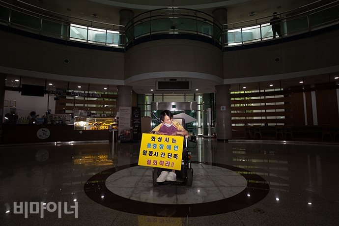 김승현(38) 씨가 기자회견이 열리기 전 화성시청 로비에서 1인 시위를 하고 있다. 목에건 손팻말에는 ‘화성시는 중증장애인 활동시간단축 철회하라’라고 적혀있다. 사진 박승원