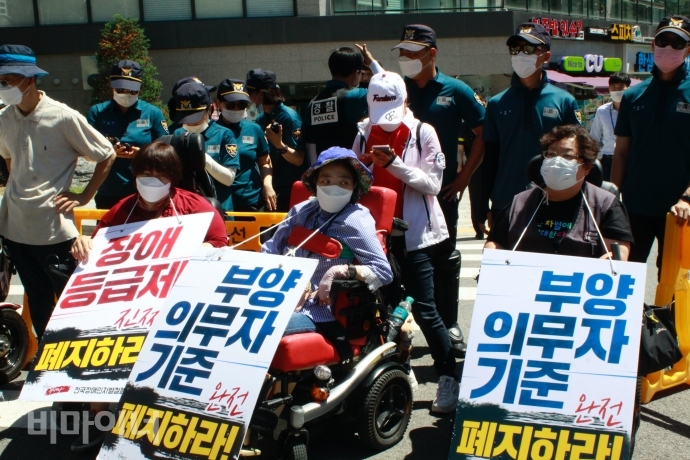 결의대회 참가자들이 피켓을 목에 걸고 경찰 앞에서 자리를 지키고 있다. ‘피켓에는 부양의무자기준 완전 폐지하라’, ‘장애등급제 진짜 폐지하라’라고 적혀있다. 사진 이가연 결의대회 참가자들이 피켓을 목에 걸고 경찰 앞에서 자리를 지키고 있다. ‘피켓에는 부양의무자기준 완전 폐지하라’, ‘장애등급제 진짜 폐지하라’라고 적혀있다. 사진 이가연
