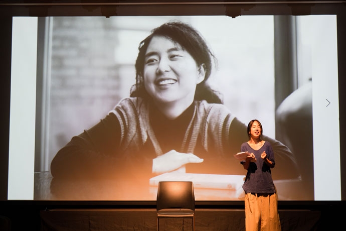 무대 중앙에 빈 의자가 있고, 그 오른편에 분홍색 통이 넓은 바지에 남색의 티셔츠를 입은 수영이 서 있다. 수영 뒤에 있는 스크린에는 긴 머리에 파마머리를 한 여성이 활짝 웃고 있는 흑백사진이 띄어져 있다. 사진 김덕중 무대 중앙에 빈 의자가 있고, 그 오른편에 분홍색 통이 넓은 바지에 남색의 티셔츠를 입은 수영이 서 있다. 수영 뒤에 있는 스크린에는 긴 머리에 파마머리를 한 여성이 활짝 웃고 있는 흑백사진이 띄어져 있다. 사진 김덕중