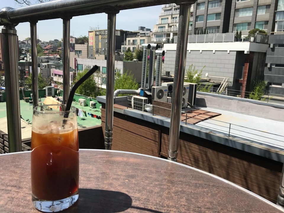 옥상 주변 풍경이 보이는 가운데 테이블 위 한 유리잔에 얼음을 띄운 차가운 커피가 놓여 있다. 내가 안전함을 느끼는 카페에서 좋아하는 커피를 마시며 반가운 사람들을 기다리는 시간. ⓒ에디