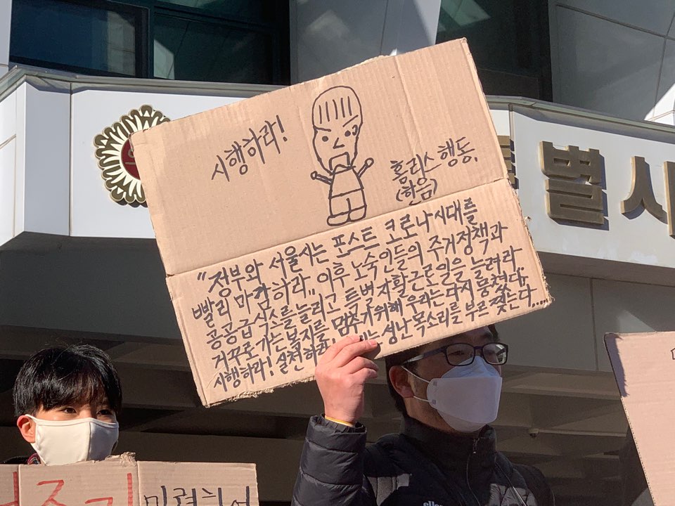 23일, 서울시의회 의원회관 앞에서 한 참가자가 "시행하라! (홈리스행동 한음) 정부와 서울시는 포스트 코로나시대를 빨리 마감하라. 이후 노숙인들의 주거정책과 공공급식소를 늘리고 특별 자활 근로일을 늘려라. 거꾸로 가는 복지를 멈추기 위해 우리는 다시 뭉쳤다. 시행하라! 실천하라! 성난 목소리를 부르짖는다."라고 적힌 피켓을 들고 있다.  사진제공 2020홈리스팀