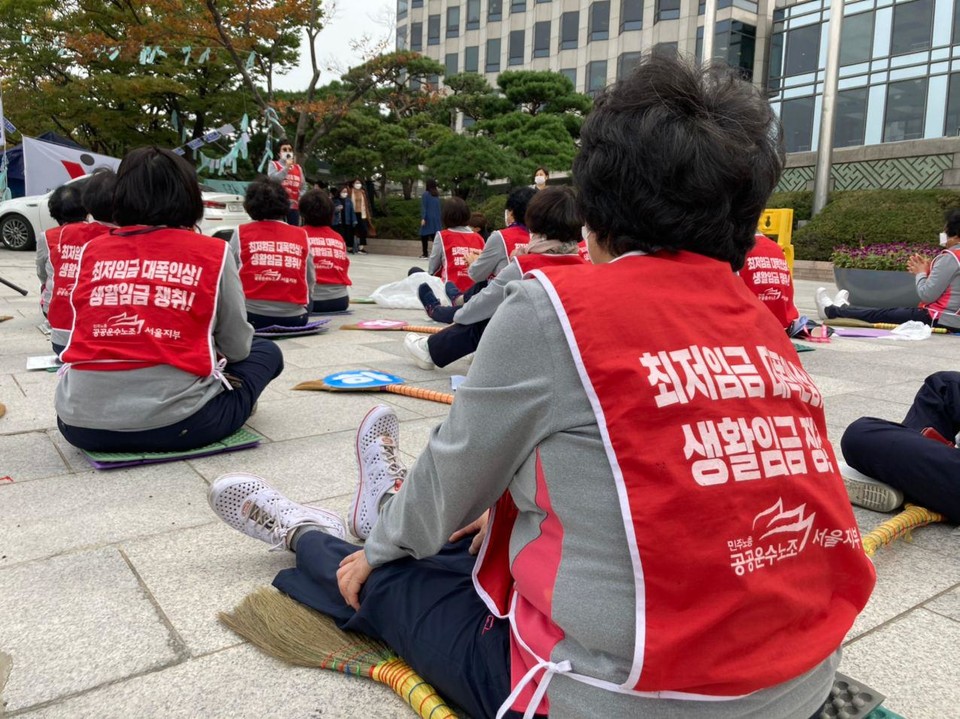 투쟁 승리를 위한 결의대회에 참석한 청소노동자들의 뒷모습. 붉은 조끼에는 “최저임금 대폭 인상! 생활임금 쟁취!”라고 쓰여 있다. 사진 황지수