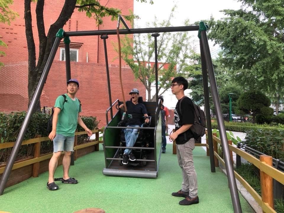 대학로 마로니에공원에 설치된 휠체어그네. 휠체어 탄 사람이 그네를 타고 있다. 그 양옆으로 비장애인이 서 있다. 사진 노들장애인자립생활센터