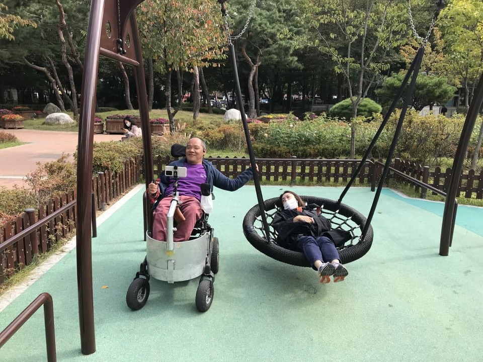 당산공원 무장애놀이터에 있는 스윙그네. 휠체어 탄 사람이 있고, 그 옆에 있는 스윙그네에 한 사람이 누워 있다. 사진 노들장애인자립생활센터