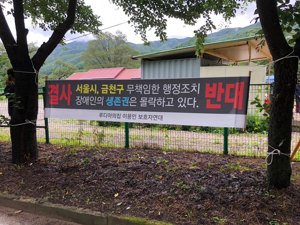 7월 13일 루디아의집 임시전원조치 당일, 정문 앞 나무에 ‘루디아의집 이용인 보호자연대’ 명의로 ‘결사반대. 서울시, 금천구 무책임한 행정조치 장애인의 생존권은 몰락하고 있다’라는 현수막이 걸려있다. ⓒ장애와인권발바닥행동