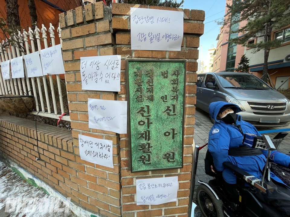 활동가들은 '집단감염 시대! 긴급탈시설 이행하라', '장애인 거주시설 폐쇄하라' 등을 적은 피켓 수십 장을 신아원 현판 주변에 붙였다. 현판 오른쪽에 장애인 활동가가 신아원 정문을 막고 있다. 사진 하민지