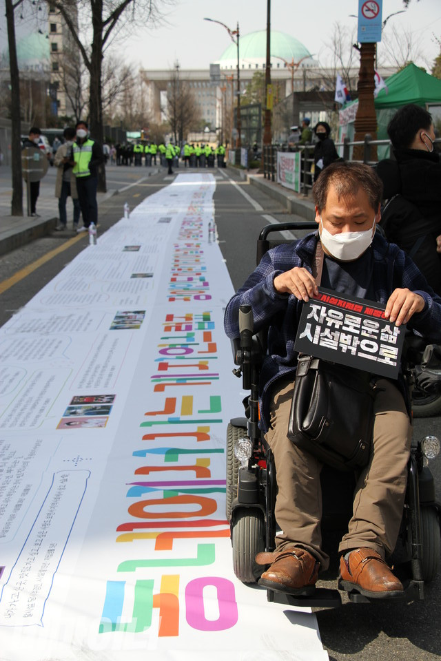 탈시설을 염원하는 탈시설장애인 100명의 편지가 60m짜리 플래카드에 적혀 이룸센터 앞 탈시설지원법·장애인권리보장법 제정 촉구 농성장을 지나 국회를 향해 깔렸다. 플래카드 옆에서 장애인 활동가가 “자유로운 삶, 시설 밖으로”라는 문구가 적힌 손피켓을 들고 있다. 사진 허현덕