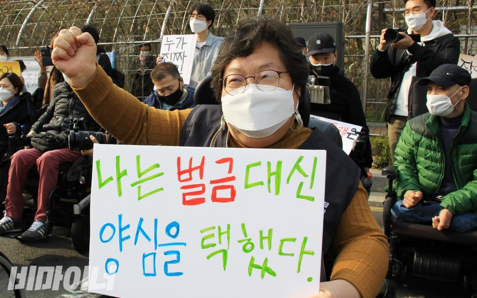 18일, 노역투쟁 기자회견에서 이형숙 서울장애인차별철폐연대 공동대표가 ‘나는 벌금 대신 양심을 택했다’라는 피켓을 들고 “투쟁!”을 외치고 있다. 사진 하민지