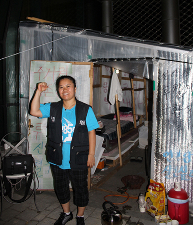 2011년 9월 5일, 용산구청 앞 신계철대위 천막. 천막 앞에서 오른쪽 주먹을 들고 투쟁을 외치는 강정희 씨가 밝은 표정으로 카메라를 바라보고 있다. ‘철인 85호 크레인’이라고 적힌 밝은 파란색 티셔츠와 검은색 조끼를 입고 있다. 사진 빈곤사회연대