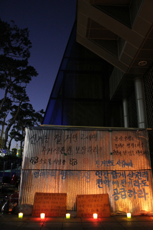 2011년 9월 5일, 용산구청 앞 신계철대위 천막. 천막에는 “신계동 철거민 2세대 주거생존권 보장하라” “소금꽃 희망꽃” “철거민도 사람이다. 인간답게 살 권리. 천막에 전기라도 공급하라” 등의 문구가 쓰여 있으며, 천막 앞에는 촛불이 놓여 있다. 사진 빈곤사회연대