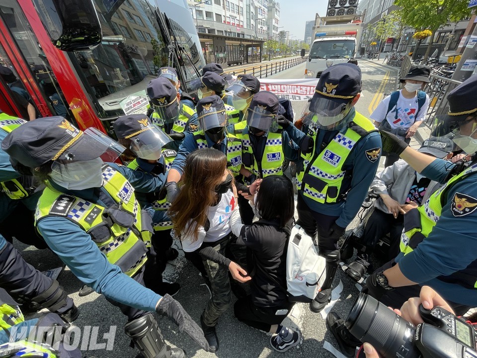 경찰 십여 명이 B3 버스 앞에 있는 두 명의 활동가를 에워싸고 내려다보고 있다. 뒤로는 다른 활동가들, 그리고 경찰의 방송 차량이 보인다. 사진 안희제