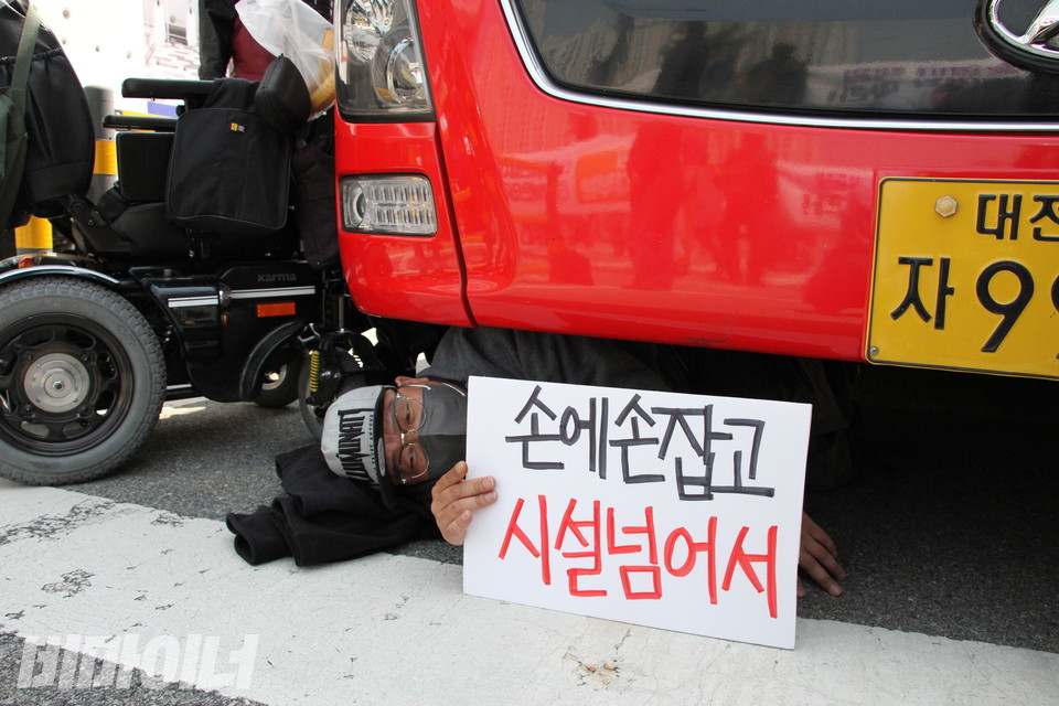 한 활동가가 B1 버스 아래에 엎드려 피켓을 들고 있다. 피켓에는 ‘손에 손잡고 시설 넘어서’라고 적혀 있고, 뒤에는 빈 휠체어가 한 대 있다. 사진 하민지