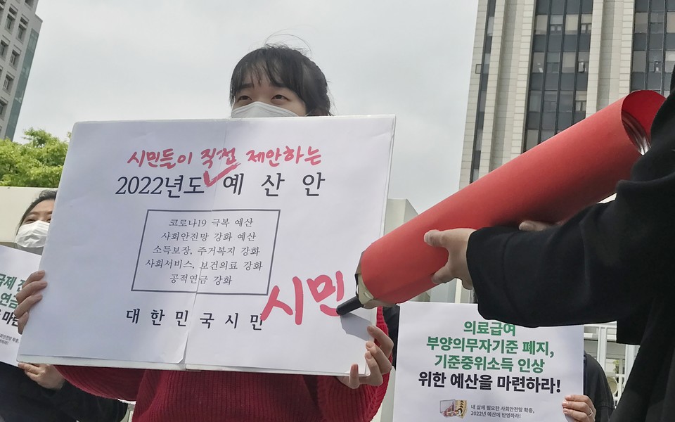 한 활동가가 피켓을 들고 있다. 피켓에는 ‘시민이 직접 제안하는 2022년도 예산안. 코로나19 극복 예산, 사회안전망 강화 예산, 소득보장, 주거복지 강화, 사회서비스, 보건의료 강화, 공적연금 강화. 대한민국 시민’이라고 적혀 있다. 다른 활동가가 피켓에 커다란 싸인펜으로 ‘시민’이라고 싸인을 하고 있다. 사진 참여연대