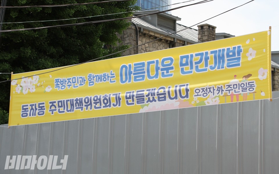 동자동쪽방촌 소유주 측이 붙인 현수막. 노란색 배경 위에 벚꽃 그림이 있다. ‘쪽방주민과 함께하는 아름다운 민간개발, 동자동 주민대책위원회가 만들겠습니다. 오정자 外 주민일동’이라고 적혀 있다. 사진 하민지