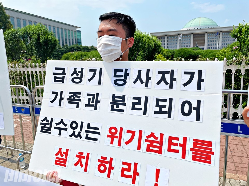 한 활동가가 국회의사당 앞에서 피켓을 들고 서 있다. 피켓에는 ‘급성기 당사자가 가족과 분리되어 쉴 수 있는 위기쉼터를 설치하라!’라고 적혀 있다. 