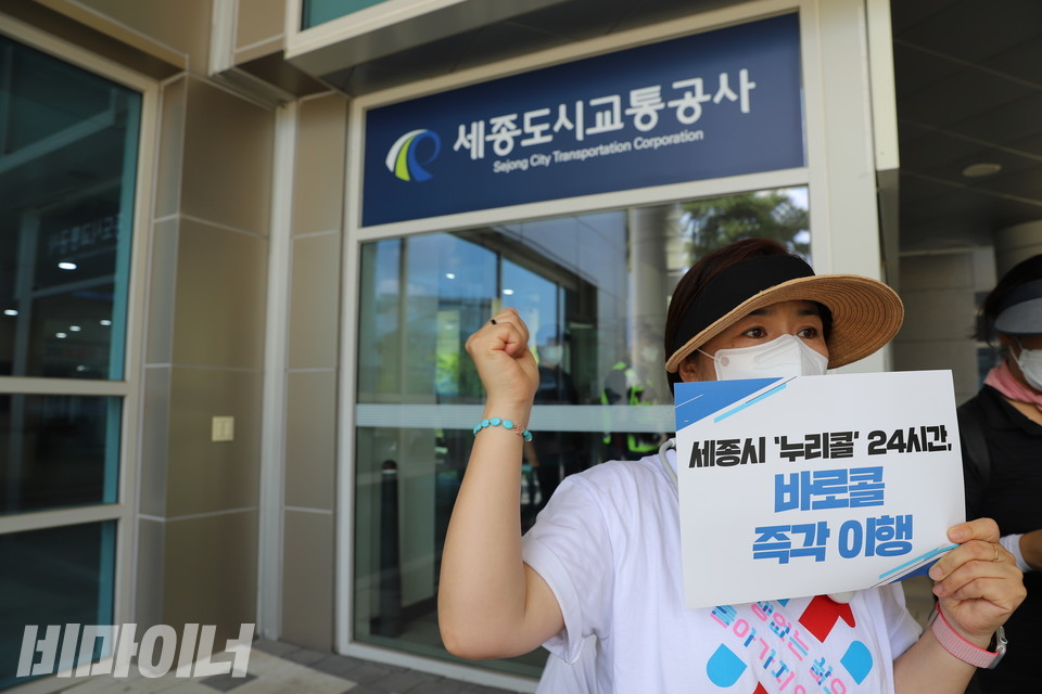 한 활동가가 ‘세종시 누리콜 24시간 바로콜 즉각 이행’이라 적힌 피켓을 들고 오른쪽 손을 들어 ‘투쟁’을 외치고 있다. 활동가 뒤로 세종도시교통공사 건물의 모습이 보인다. 사진 하민지
