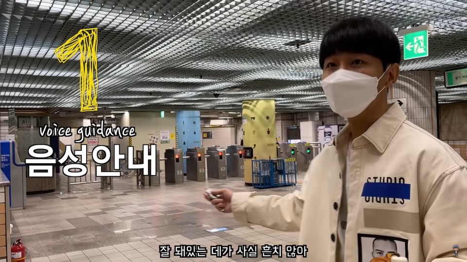 원샷한솔 ‘스크린도어 없는 지하철에서 생긴 일 | 사회실험, 실험카메라’ 유튜브 영상 캡처. ‘음성안내’라는 글자가 크게 적혀 있고, 원샷한솔이 시각장애인용 공용리모컨을 누르며 ‘잘 돼 있는 데가 사실 흔치 않아’라고 말하고 있다. 그는 장애인이 일상생활에서 겪는 어려움을 사회 구조적 문제와 연결하여 보여주고 있다.