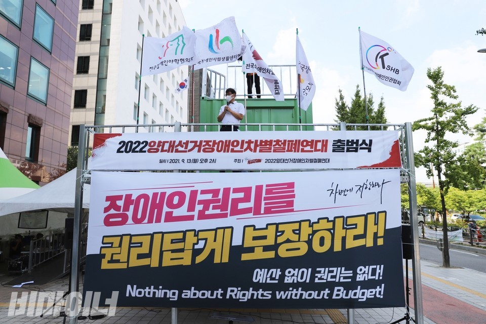 장애인탈시설지원법·권리보장법 제정투쟁 농성장. 양대선거장차연 출범식이 열리고 있다. 커다란 현수막에 ‘장애인권리를 권리답게 보장하라! 예산 없이 권리는 없다’라고 적혀 있다. 컨테이너 농성장 위에 윤종술 전국장애인부모연대 회장이 올라가 있다. 사진 하민지