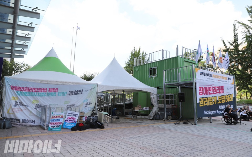 전체 농성장의 모습. 천막 농성장 두 채 옆에 2층으로 구성된 컨테이너 농성장이 있다. 농성장 앞에서 탈시설장애인당 투쟁캠프 개소식이 열리고 있다. 사진 하민지