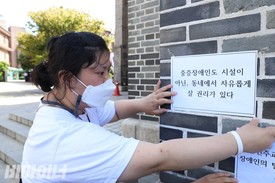 박경인 활동가가 명동성당 외벽에 피켓을 붙이고 있다. 피켓에는 ‘중증장애인도 시설이 아닌, 동네에서 자유롭게 살 권리가 있다’라고 적혀 있다. 사진 하민지