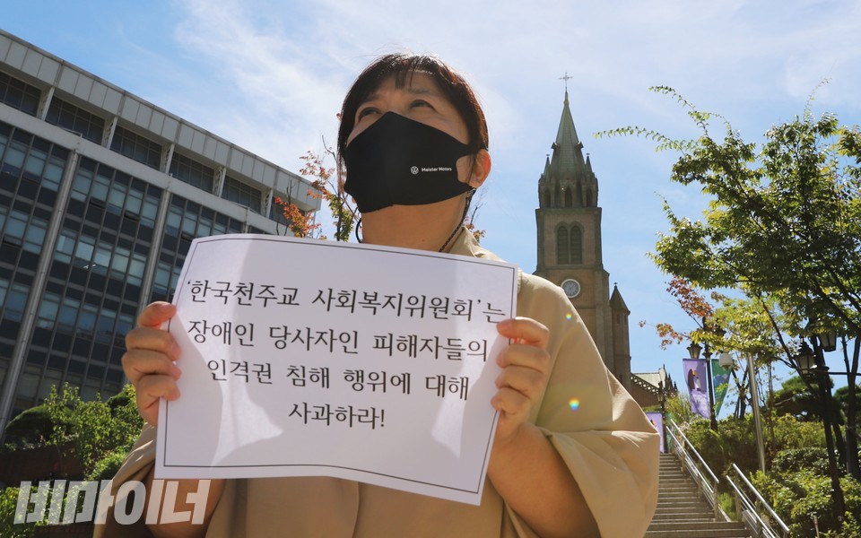 김수정 대표가 명동성당 앞에서 피켓을 들고 있다. 피켓에는 ‘한국천주교주교회 사회복지위원회는 장애인당사자인 피해자들의 인격권 침해행위에 대해 사과하라’라고 적혀 있다. 사진 하민지