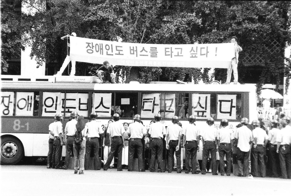 2001년 8월 29일, 서울 세종문화회관 앞에서 버스를 점거한 장애인 활동가들. 버스 위에는 “장애인도 버스를 타고 싶다!”고 적힌 현수막이 펼쳐져 있고, 버스 창문에도 “장애인도 버스 타고 싶다”는 글자가 커다랗게 적혀 있다. ⓒ장애인이동권연대 