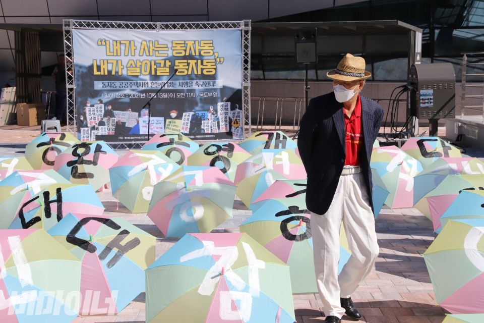 한 동자동 주민이 각종 구호가 적혀있는 우산들 사이로 뒷짐을 지고 있고, 왼편으로 "내가 사는 동자동 내가 살아갈 동자동"이라고 적힌 현수막이 보인다. 사진 이가연