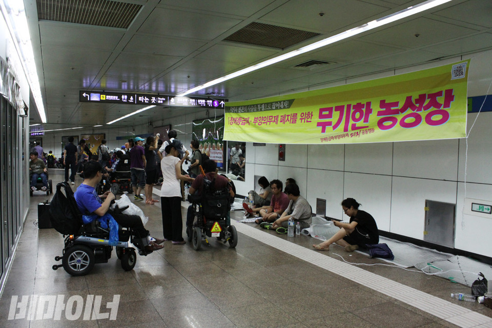 2012년 8월 21일 밤, 농성 첫날의 모습. 지붕조차 없는 바닥에 은박지 롤을 깔고 사람들이 앉아 있다. 천장에는 “장애등급제·부양의무제 폐지를 위한 무기한 농성”이라는 노란색 현수막 하나가 걸려 있다. 사진 비마이너DB