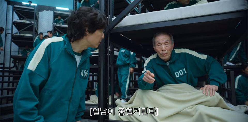 001이라는 숫자가 적힌 탁한 녹색의 츄리닝을 입고 침대에 앉아 손가락으로 앞을 가리키고 있는 일남을 456이라는 숫자가 적힌 같은 옷을 입은 기훈이 바로 앞에서 바라본다. 화면 하단에는 [일남이 중얼거린다]라는 폐쇄자막이 있다. 넷플릭스 화면 캡쳐.
