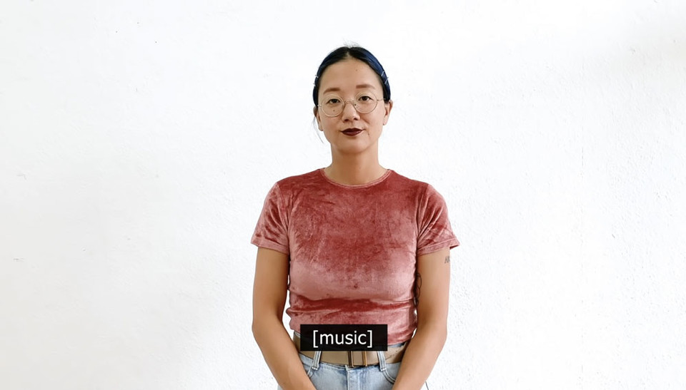 울긋불긋한 티셔츠에 청바지를 입고 테가 가는 안경을 쓴 크리스틴 선 킴이 무뚝뚝한 표정으로 서 있다. [music]이라는 짧은 폐쇄자막이 떠 있다. 유튜브 영상 “Artist Christine Sun Kim Rewrites Closed Captions | Pop-Up Magazine” 화면 캡쳐.