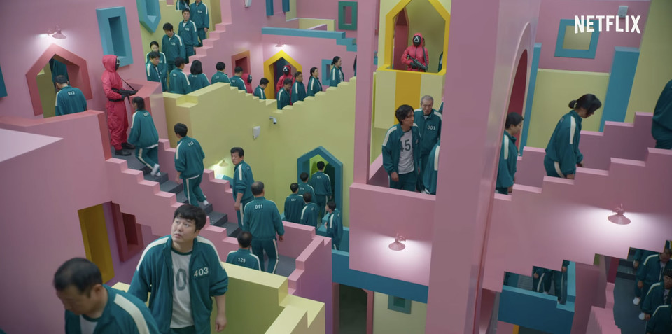 부드러운 파스텔 톤의 분홍색, 연두색, 하늘색으로 이루어진 장난감 집 같은 계단과 문들이 사방팔방에 이어져 있고, 어느 계단도 그 목적지가 드러나지 않는다. 소수의 관리자가 총을 들고 감시하는 가운데 참가자들은 이 계단을 일렬로 따라가고, 몇 참가자는 고개를 들어 크게 뜬 눈으로 주변을 살핀다. ‘오징어 게임’ 넷플릭스 공식 예고편 중.