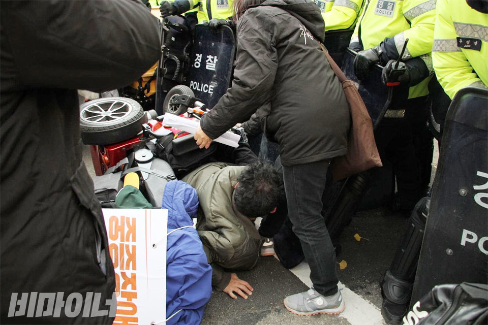 경찰의 과잉진압으로 인천 민들레장애인자립생활센터에서 활동하는 유재근 활동가가 휠체어를 탄 상태로 옆으로 쓰러졌다. 휠체어에 깔린 유 활동가가 손을 바닥에 짚으며 일어나고 있고 다른 활동가가 이를 돕고 있다. 사진 강혜민