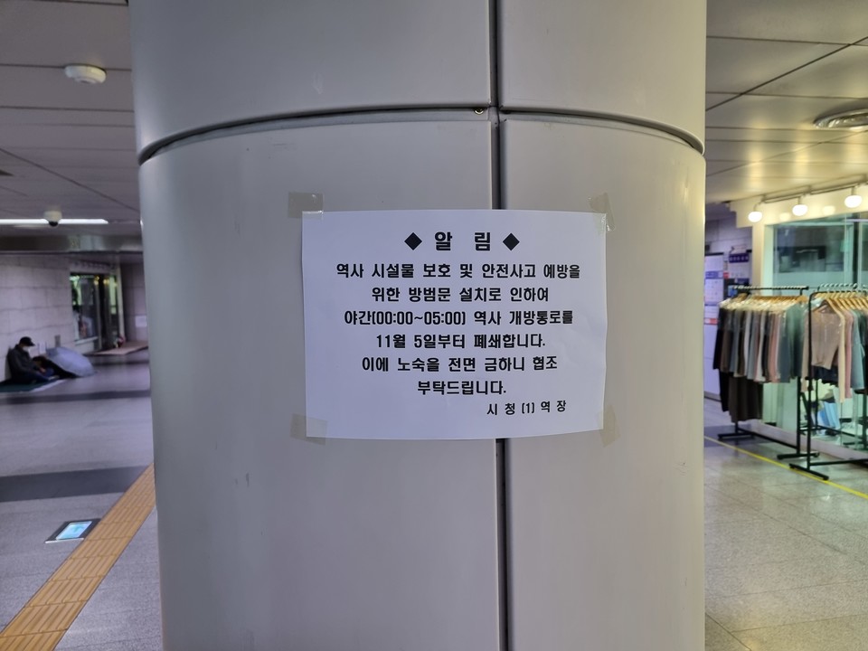 11월 4일 21시에 촬영한 시청역 1-6-7번 출구 사이 지하도 폐쇄 조치에 관한 안내문. 안내문에는 다음과 같이 적혀 있다. "역사 시설물 보호 및 안전사고 예방을 위한 방범문 설치로 인하여 야간 시간인 0시부터 오전 5시까지 역사 개방통로를 11월 5일부터 폐쇄합니다. 이에 노숙을 전면 금하니 협조 부탁드립니다. 시청역장." 안내문이 붙은 기둥 양옆으로 좌측에는 자리를 펴고 앉아 있는 거리홈리스 당사자가 있고, 우측에는 불을 밝힌 옷가게가 있다. 제공 주장욱 노숙인인권공동실천단 활동가